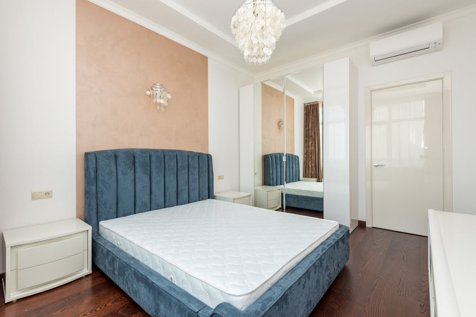 A modern bedroom with a white upholstered bed featuring a high, vertically tufted headboard, positioned next to a small white bedside table with a decorative vent. The room has plain white walls, a da